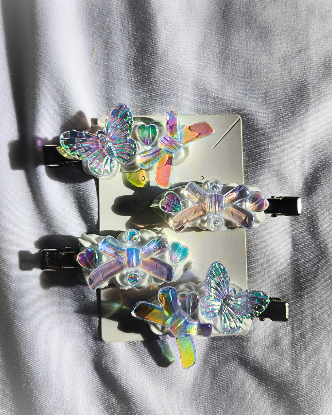 Four butterfly-shaped hair clips with iridescent holographic wings and black metal clasps, arranged on a white card over a light gray fabric background.