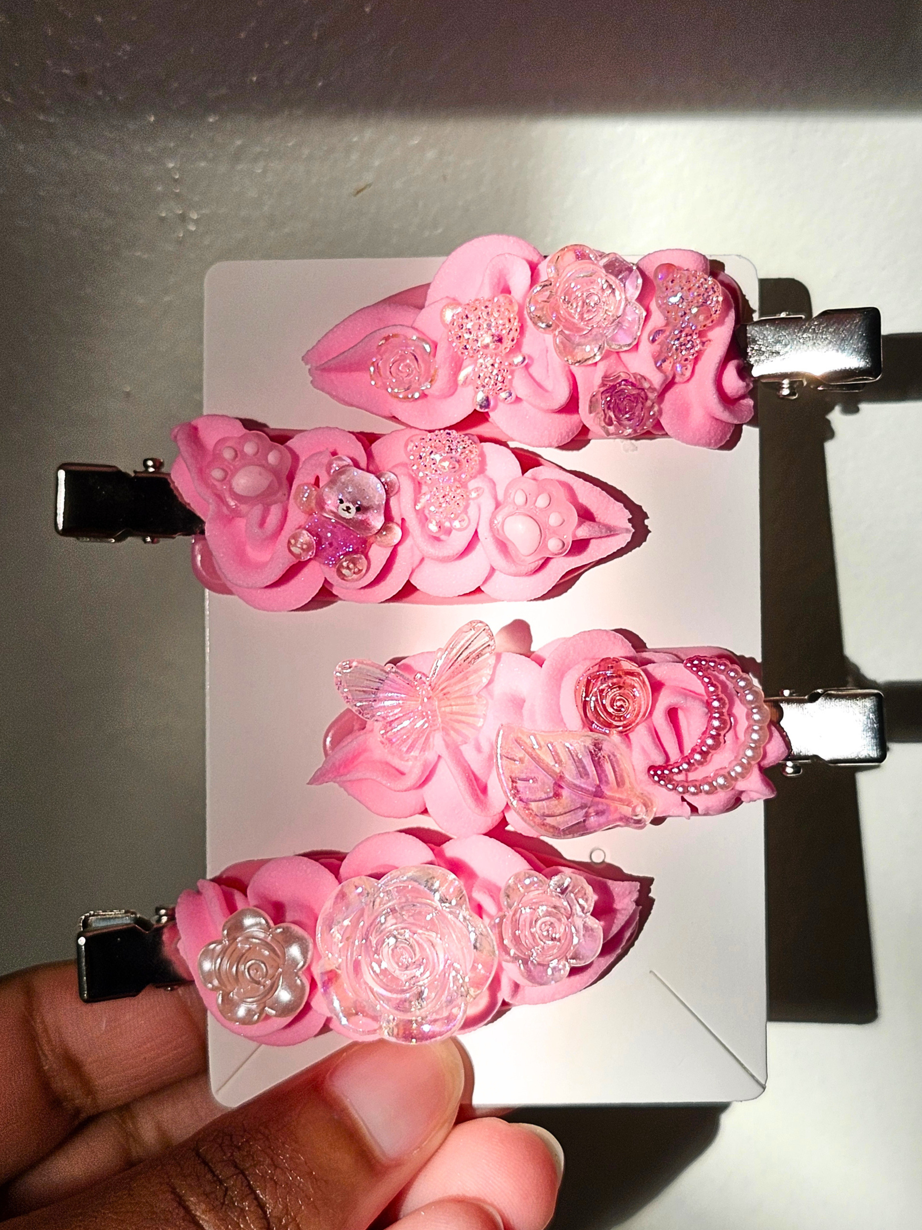 Four pink decorative hair clips with floral and butterfly embellishments, displayed on a white card with a hand holding the bottom corner.