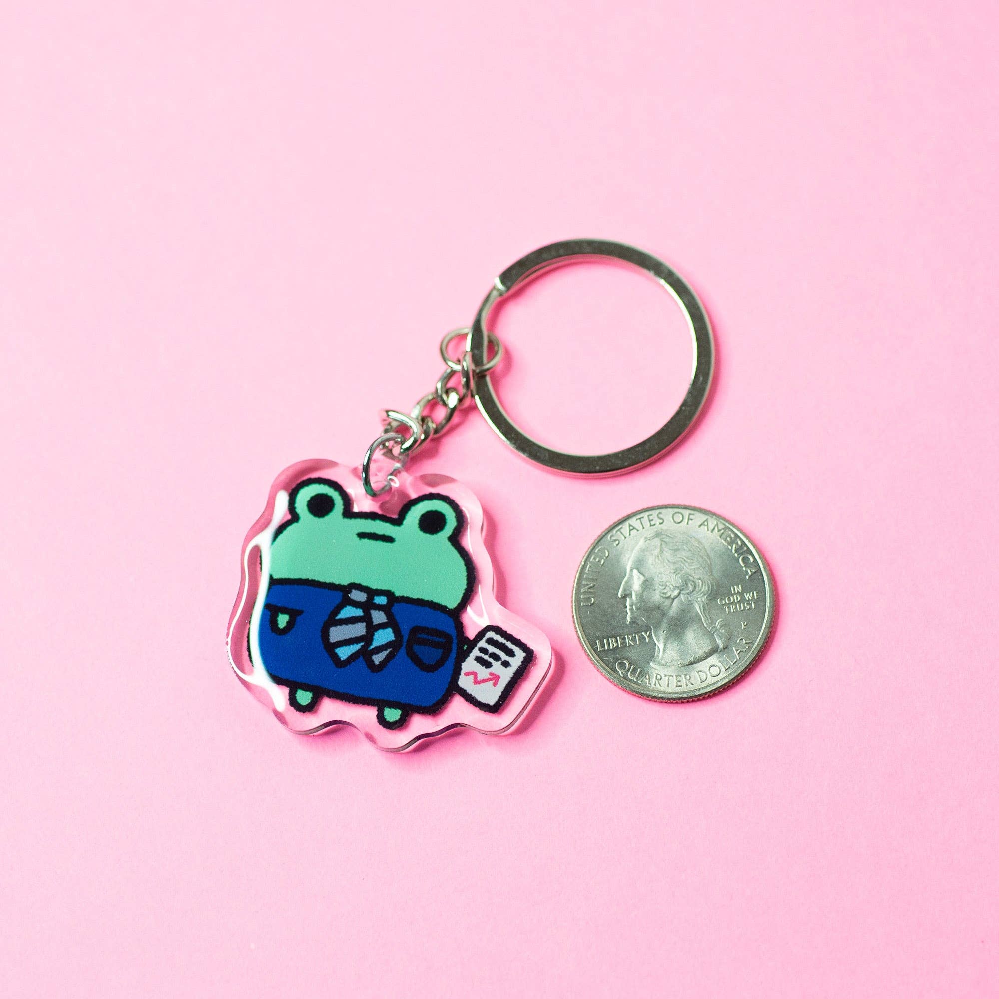 Keychain with cartoon frog design next to a quarter on a pink background