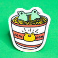 Illustrated sticker of a cartoon character eating noodles from a cup with 'nom nom' text on a green background
