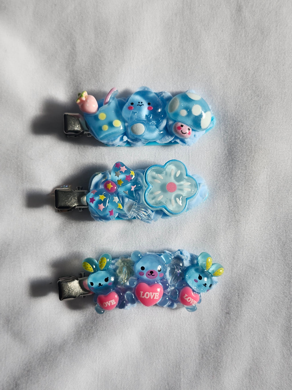 Three small blue hair clips with cute cartoon bear and flower charms, arranged vertically on a white textured fabric background.