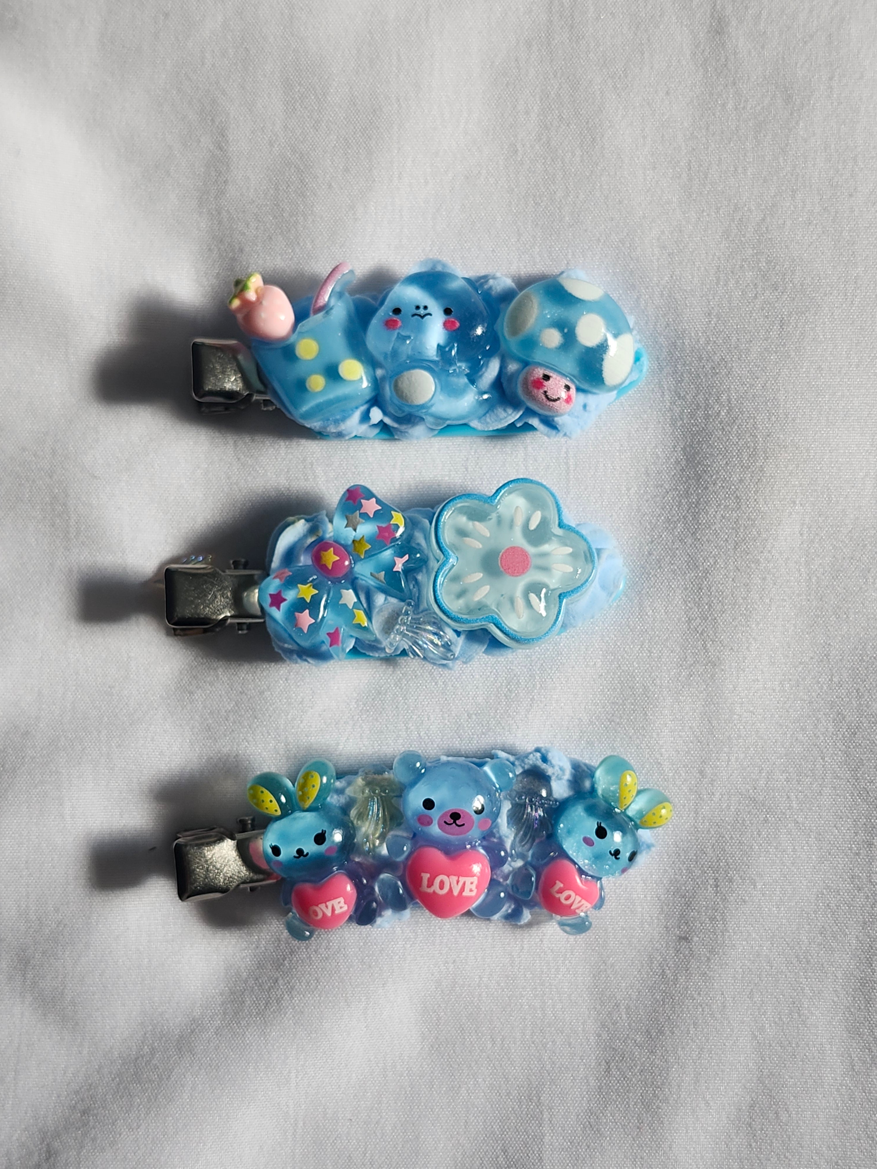 Three small blue hair clips with cute cartoon bear and flower charms, arranged vertically on a white textured fabric background.