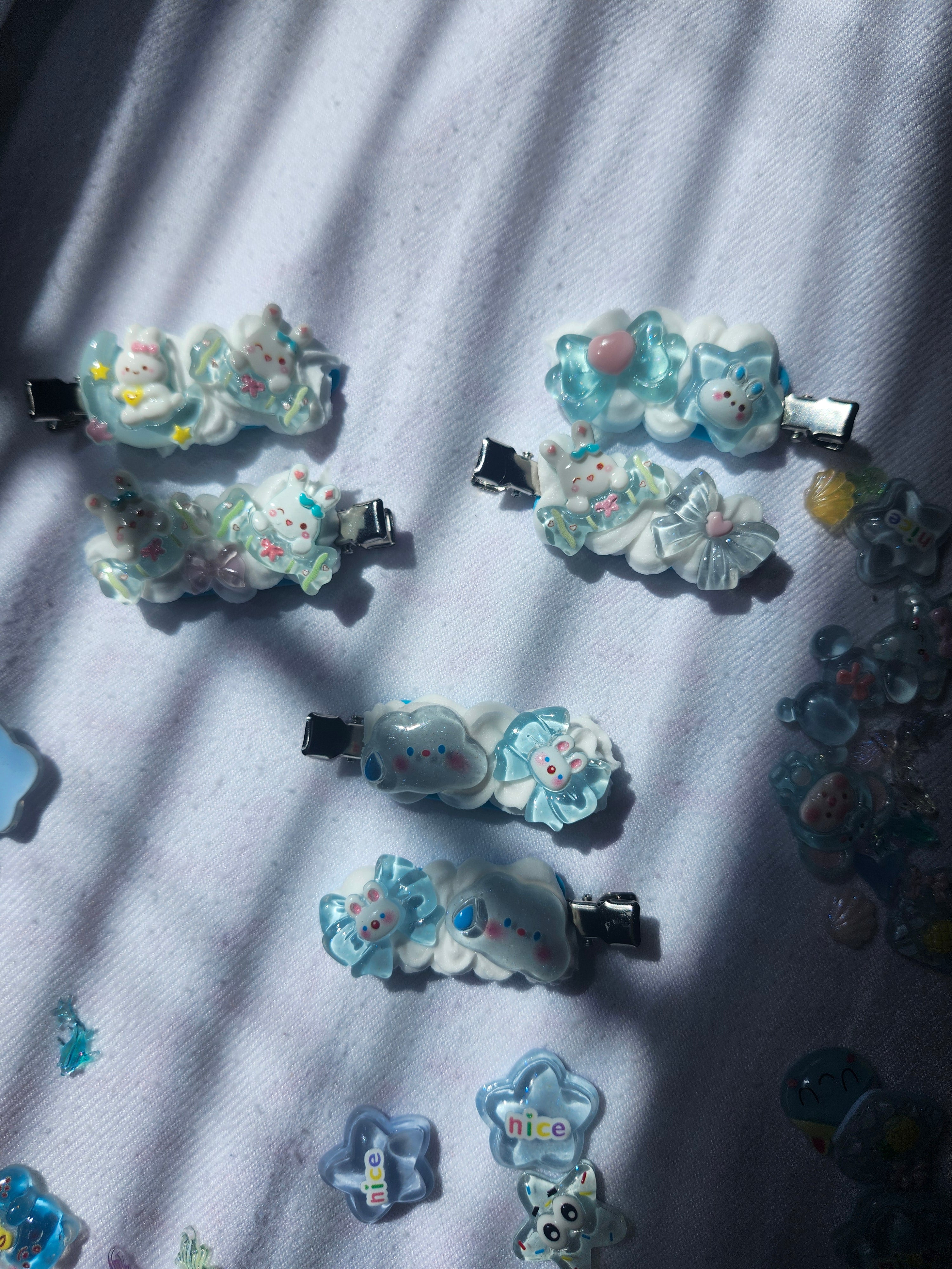 Several small decorative hair clips with pastel blue and white cartoon animal and flower charms are arranged on a white fabric surface with strong diagonal shadows. Some loose matching charms are scattered around the edges.