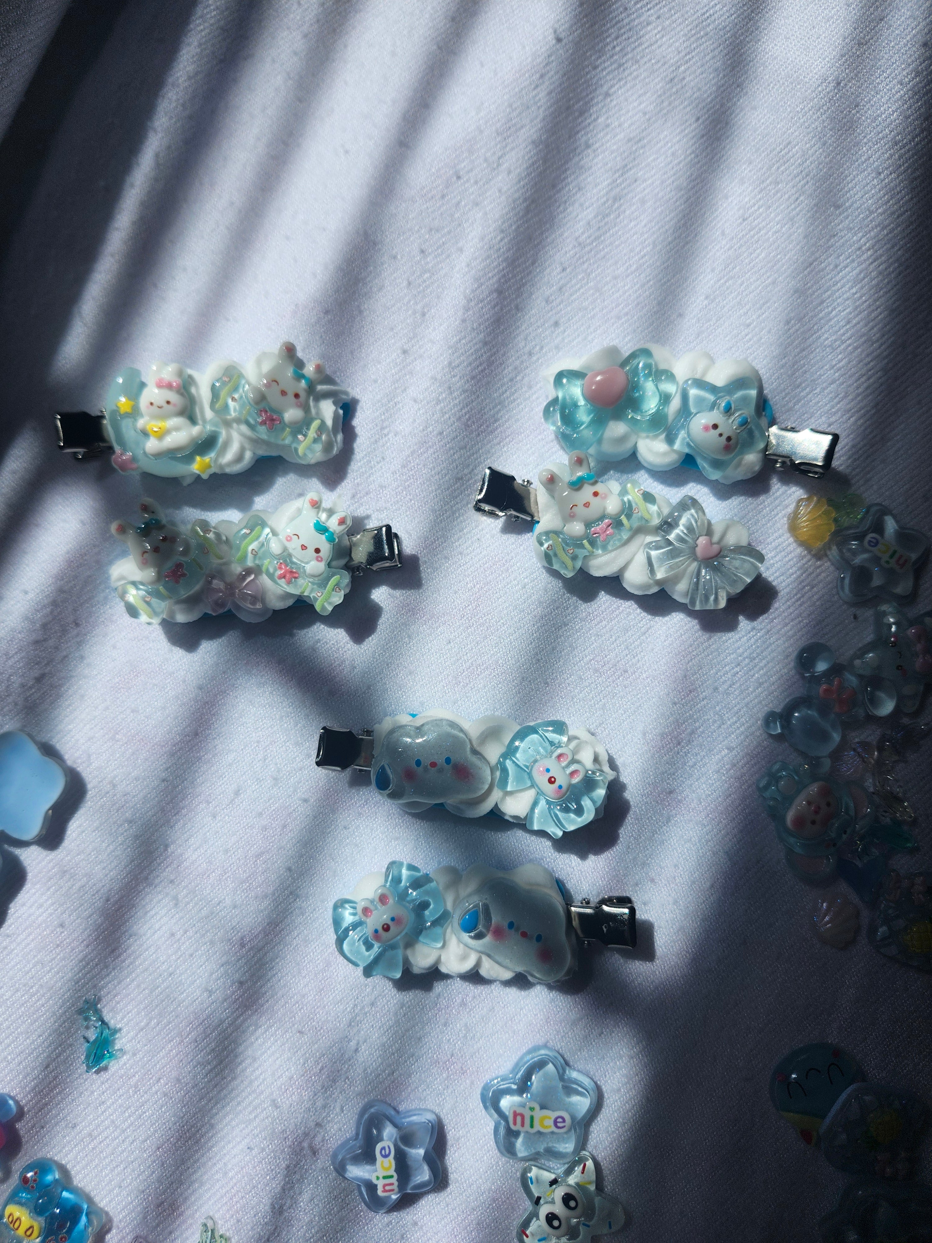 Several small decorative hair clips with pastel blue, white, and mint charms are arranged on a white fabric surface with strong diagonal shadows. The clips feature floral and bunny-style embellishments in soft pastel colors, and some translucent blue charms are also visible nearby.