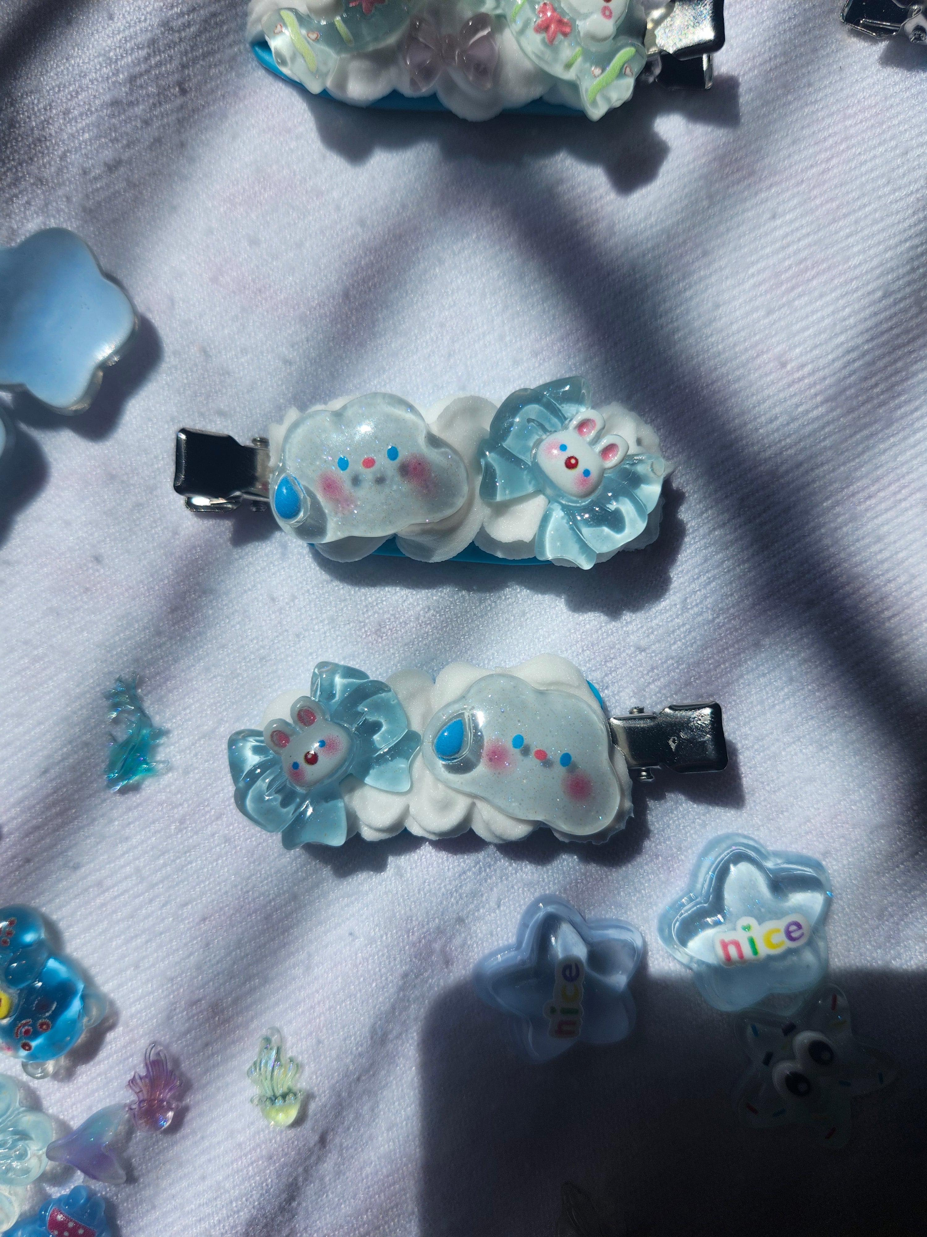 two small hair clips with cute bunny and cloud designs in light blue, white, and pink, arranged on a white fabric background with some loose decorative accessories nearby.