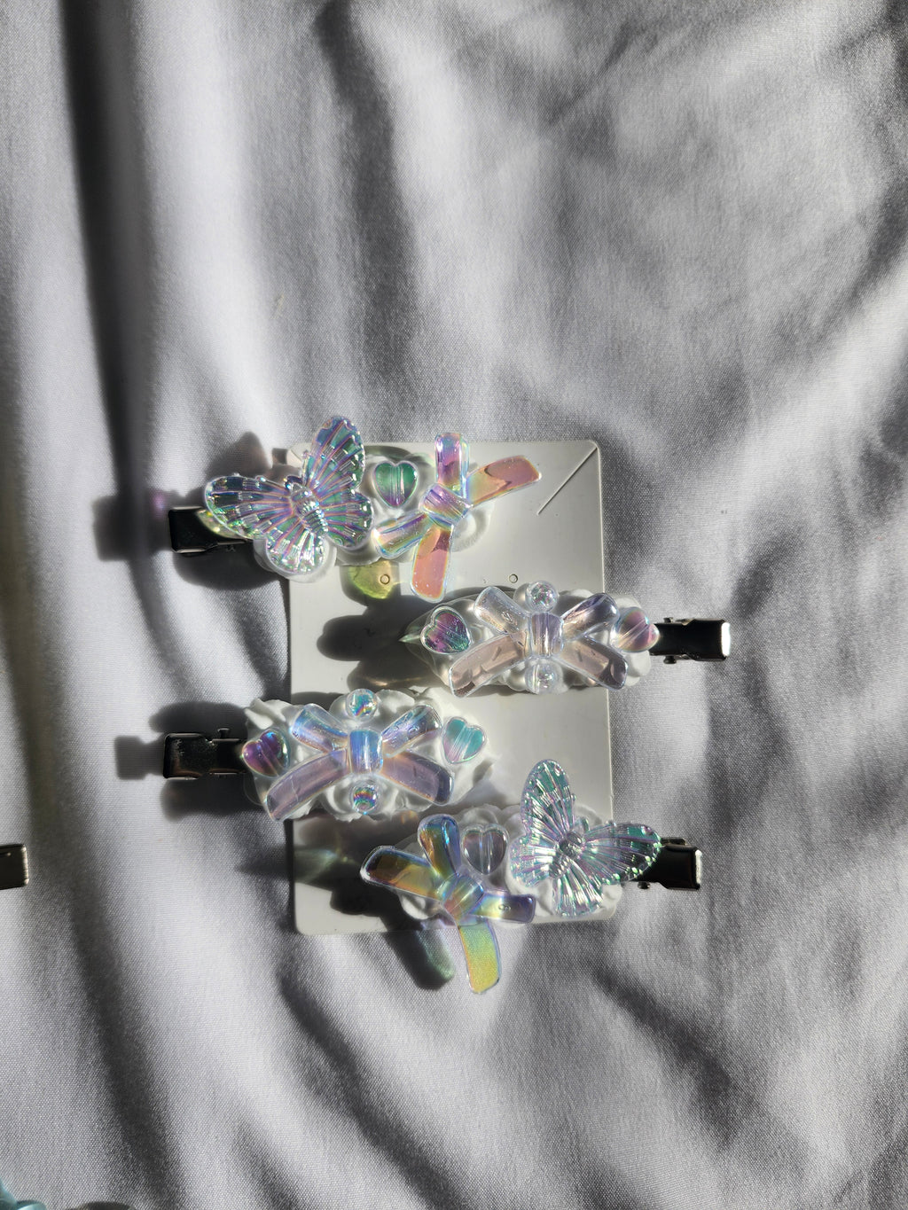 Four butterfly-shaped hair clips with iridescent holographic resin wings and black metal clips, arranged on a white card over a white fabric background.