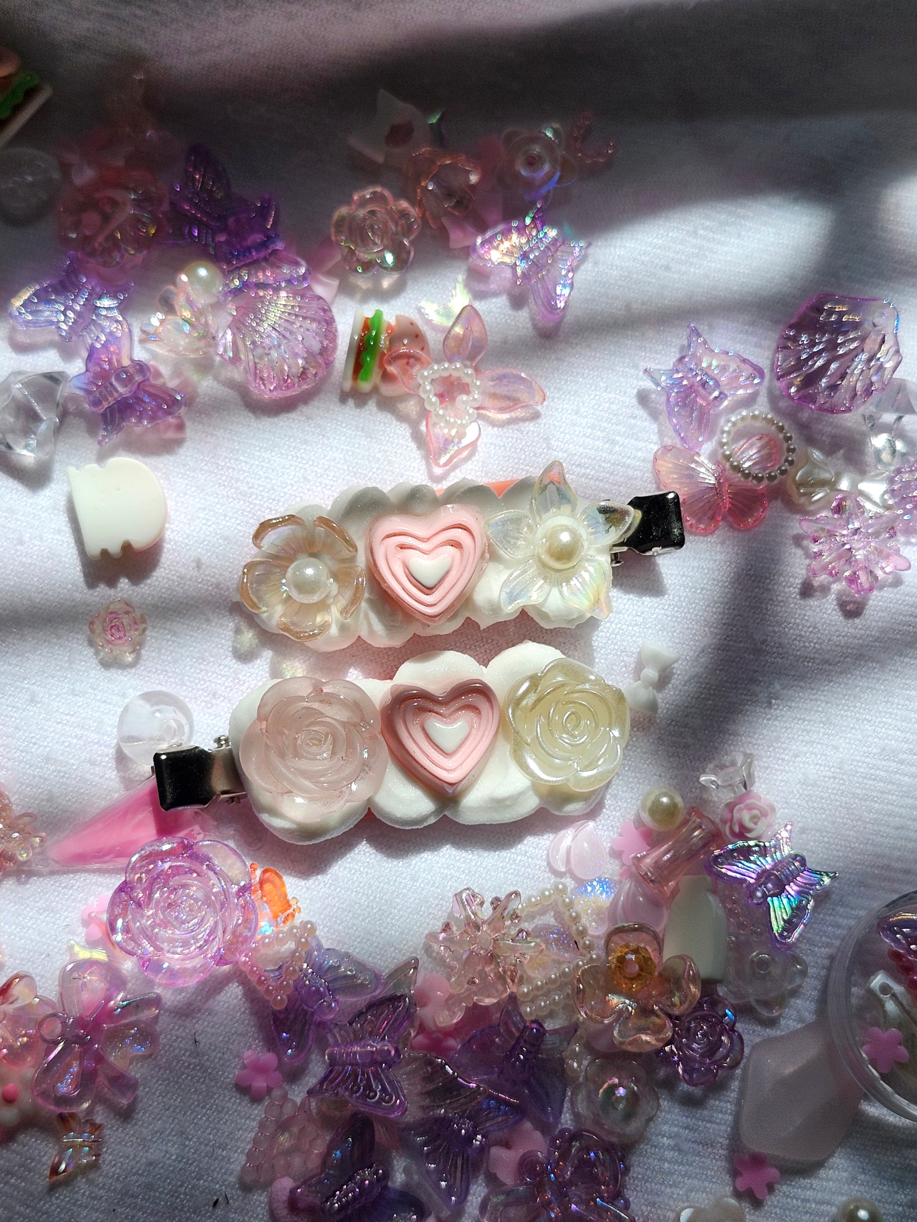 Two pink and cream hair clips decorated with faux flowers, pearls, and heart-shaped beads, surrounded by scattered pink and purple craft beads on a white fabric background.