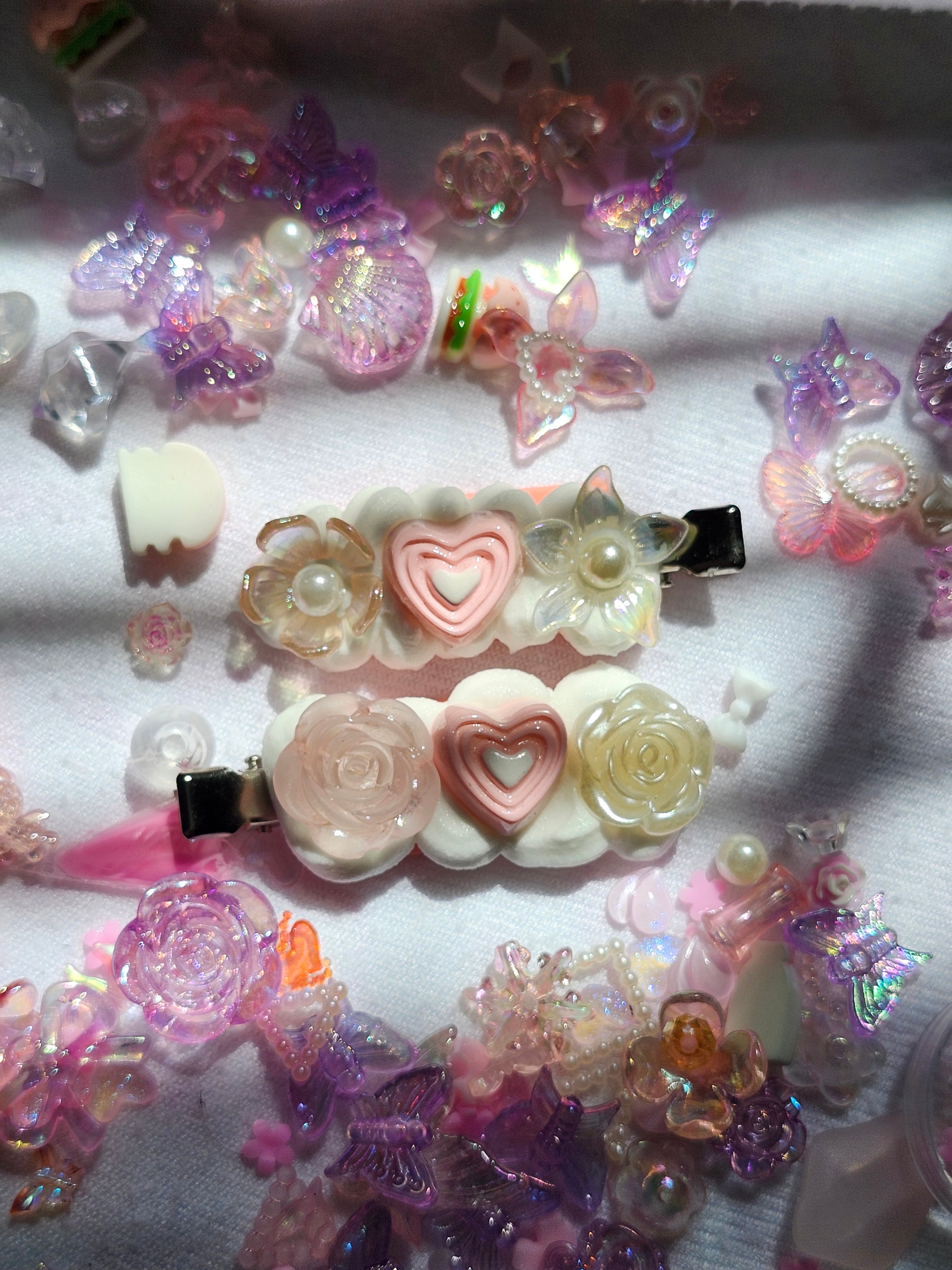 Two decorative hair clips with pastel pink and cream floral and heart designs, surrounded by scattered matching pink and purple plastic charms, beads, and butterfly embellishments on a white fabric background.